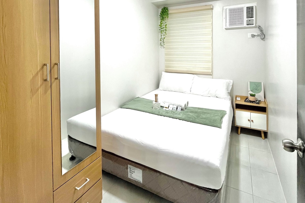 Un dormitorio con cama, armario, mesita de noche, espejo, ventana con persianas, unidad de aire acondicionado y una planta pequeña.