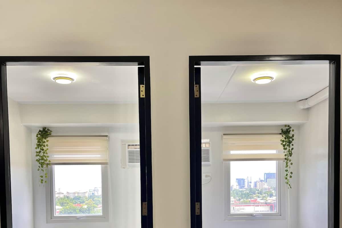 Reflejo de un cuarto de hotel con cama, almohadas, cabecera de madera, ventana con persianas y vista panorámica de la ciudad.