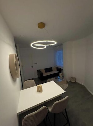 Ein moderner Essbereich mit einem weißen Tisch, zwei Stühlen, einer außergewöhnlichen runden Leuchte und einer Couch im Hintergrund.