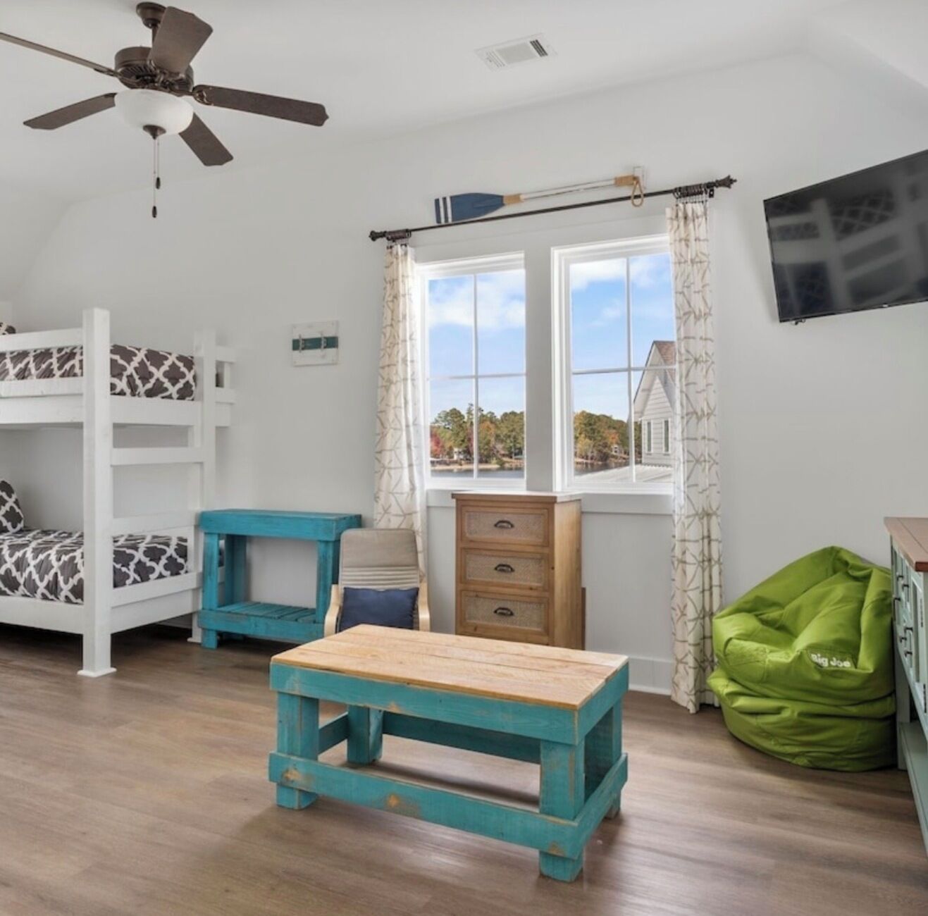Quarto com beliches, uma mesa de madeira, um pufe verde, uma janela com cortinas e um ventilador de teto.