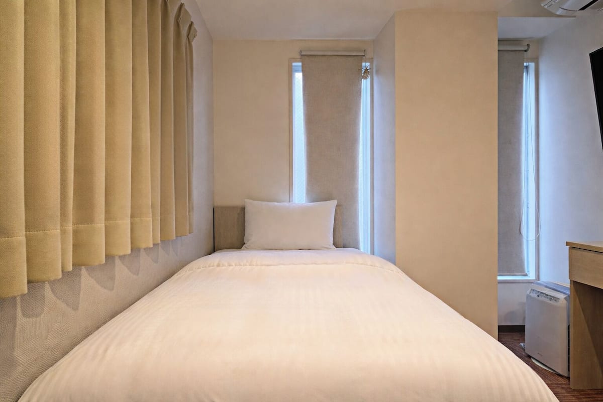 A single bed with a headboard, beige curtains, and a window with a view.