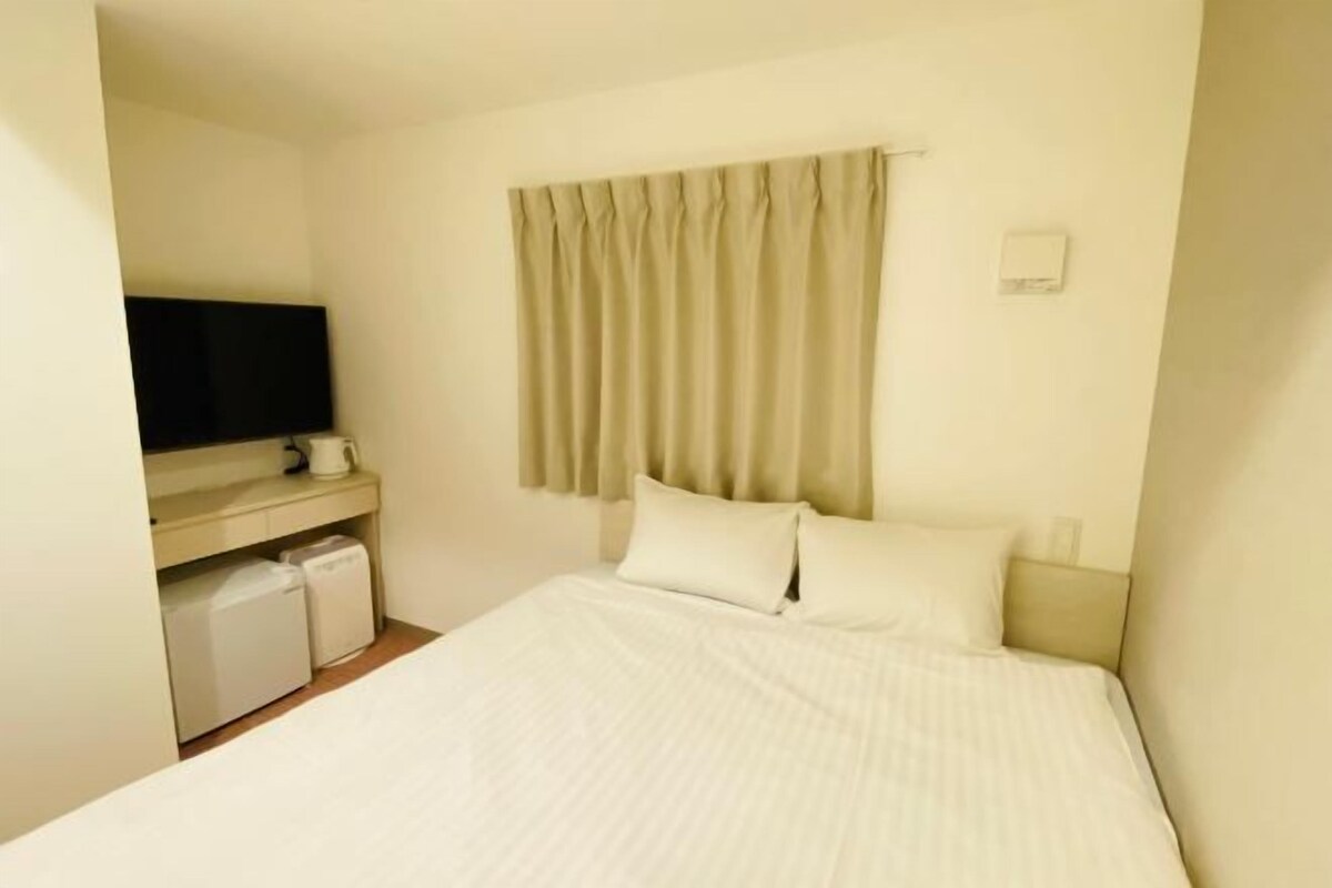 A hotel room with a bed, a television on a stand, a small desk, and a window with curtains.