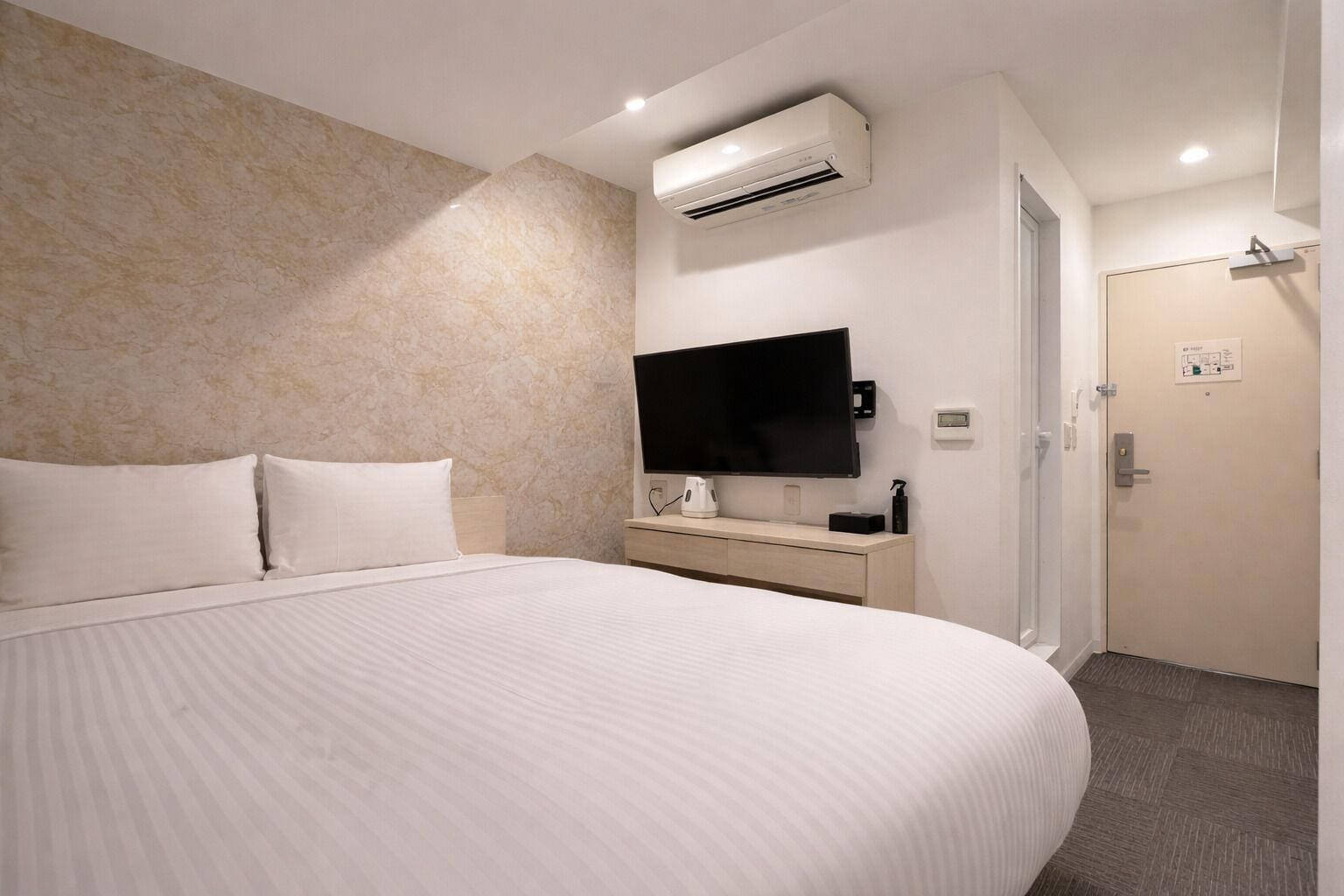 A hotel room with a large bed, a flat-screen TV, an air conditioning unit, a bedside table with a lamp, and a door with a lock.