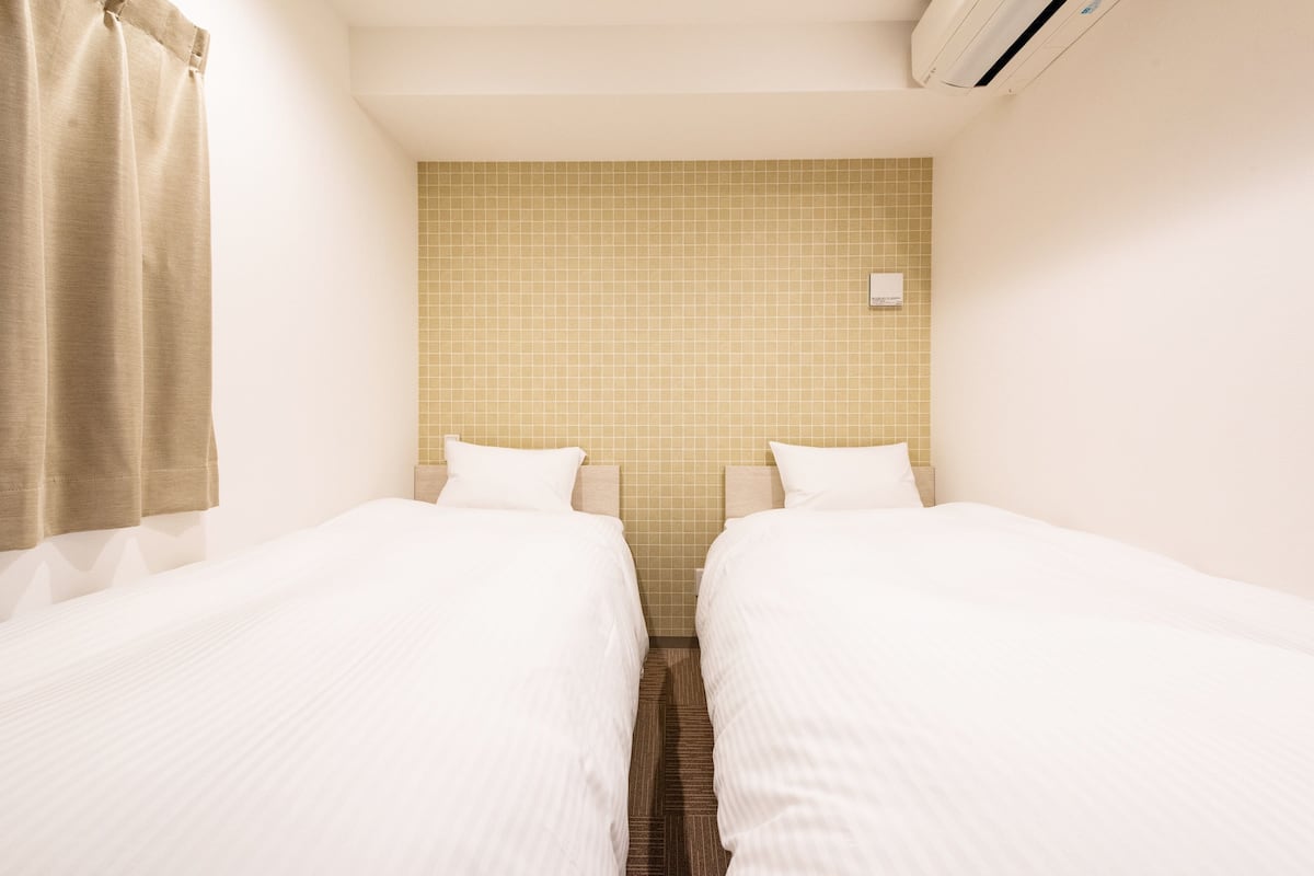 A hotel room with two single beds, a shower curtain, a tiled wall, and an air conditioning unit.