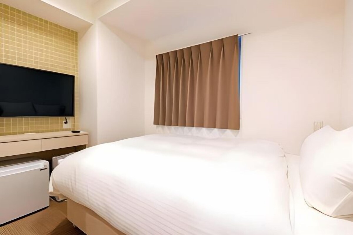 A hotel room with a bed, a small bedside table, a wall-mounted TV, and a window with curtains.