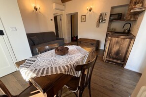Kodikas olohuone, jossa on puinen ruokapöytä, virkattu pöytäliina, ruskea nahkasohva sekä maalaishenkinen keittiö, jossa on mikroaaltouuni ja puiset kaapit.