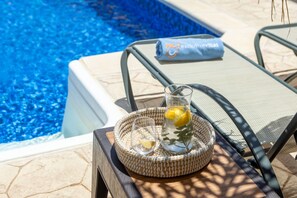 Une scène au bord de la piscine, avec un plateau en osier contenant un verre d’eau et des tranches de citron, une serviette ornée d’un logo et une chaise longue.