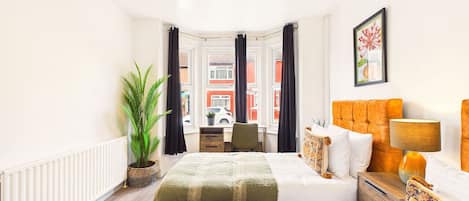 Un dormitorio con cama, mesita de noche, lámpara y vista a la calle a través de una ventana.
