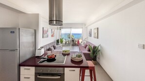 Eine kompakte Küche mit Edelstahl-Dunstabzugshaube, eingebautem Backofen und Kühlschrank. Ein Essbereich mit Tisch und Stühlen bietet Blick auf das Meer durch eine Glastür.