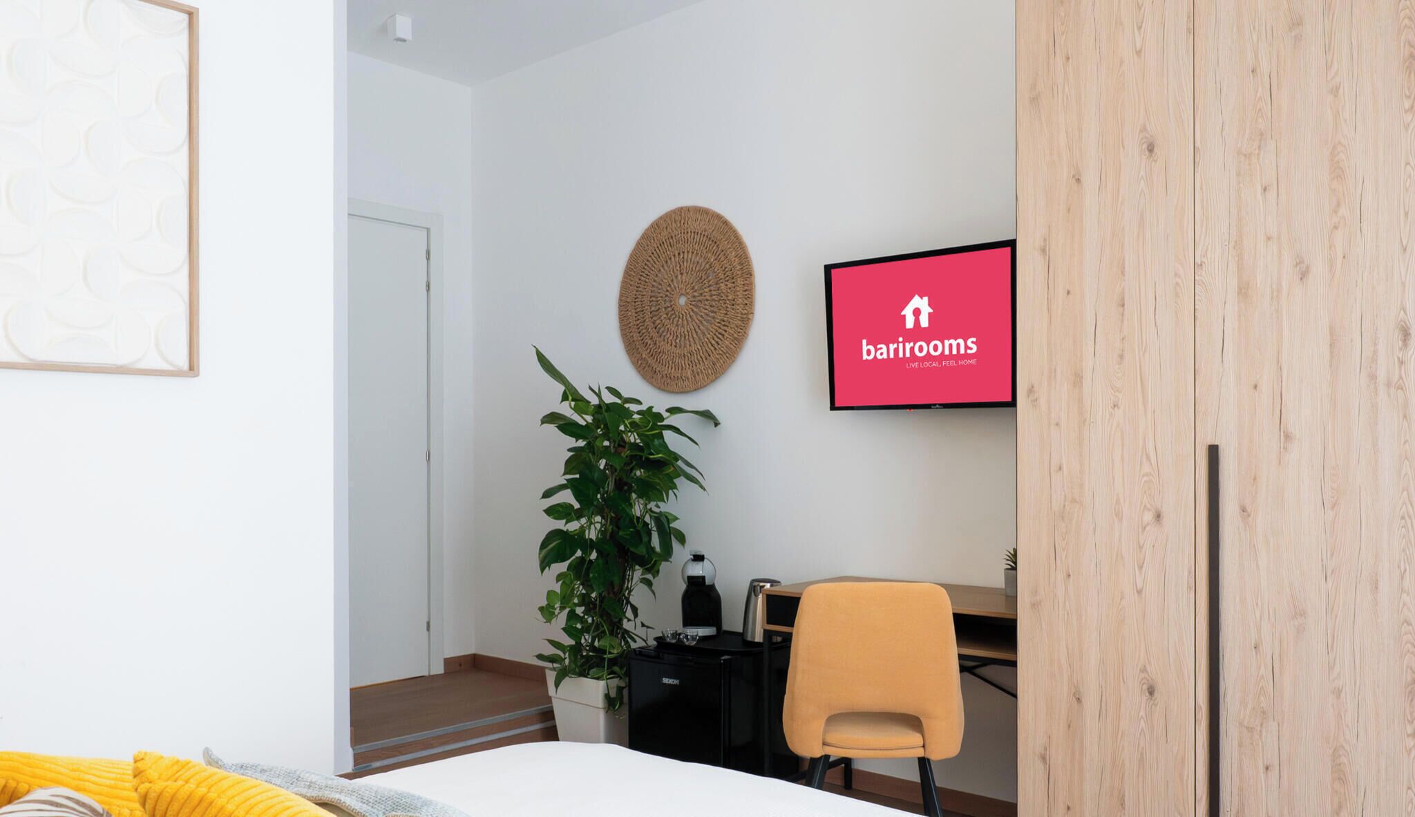 Une chambre d’hôtel moderne équipée d’un lit, d’un bureau, d’une chaise, d’une plante en pot et d’un téléviseur fixé au mur, sur lequel est affiché le logo de Barriorms.
