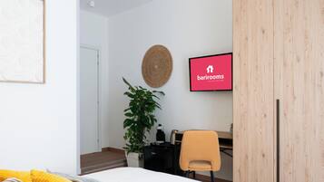 Une chambre d’hôtel moderne équipée d’un lit, d’un bureau, d’une chaise, d’une plante en pot et d’un téléviseur fixé au mur, sur lequel est affiché le logo de Barriorms.