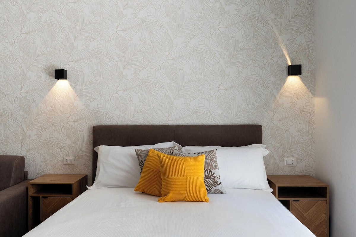 Una cama bien hecha con edredón blanco, dos mesitas de noche, un cabecero marrón y una pared con papel tapiz estampado con hojas.