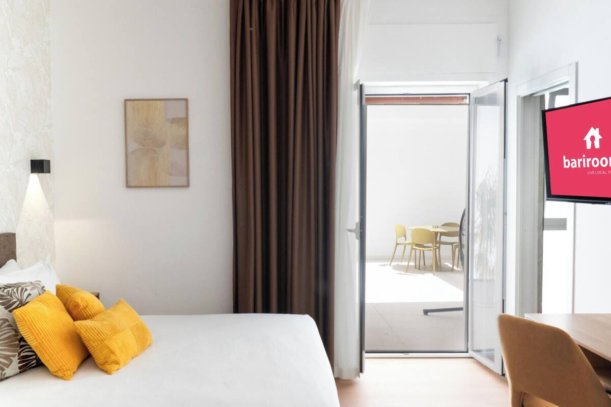 Habitación de hotel con cama, escritorio, silla, televisor y puerta corrediza de vidrio que da a un balcón.