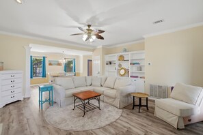 Ein Wohnzimmer mit Ecksofa, Couchtisch, Beistelltisch, Deckenventilator und Bücherregal.