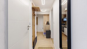 Un pasillo estrecho con una puerta de madera, una pequeña cocina con microondas y un cubo de basura.