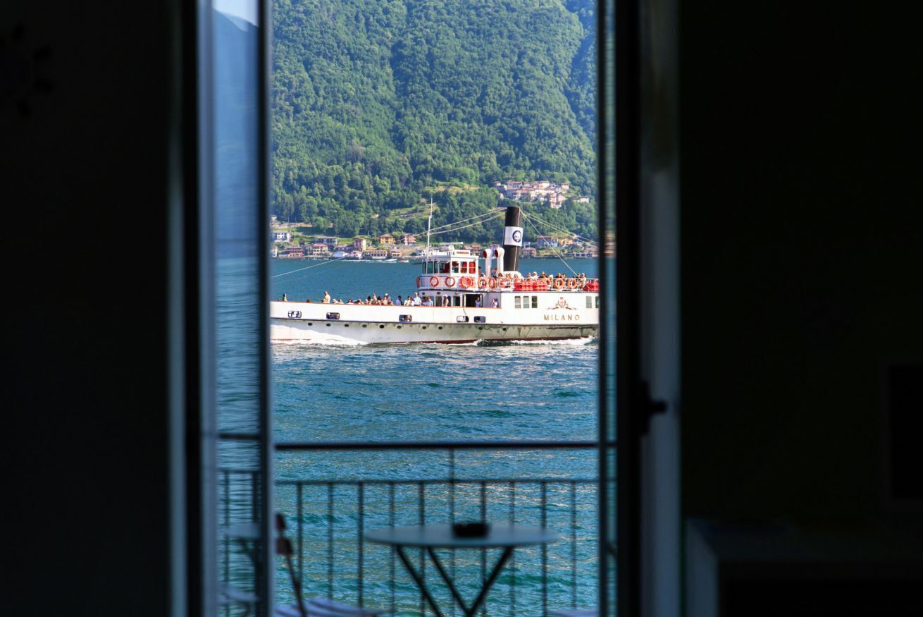 A view from a balcony with a table and railing, overlooking a body of water with a boat named 'MILANO' and a mountainous landscape in the background.