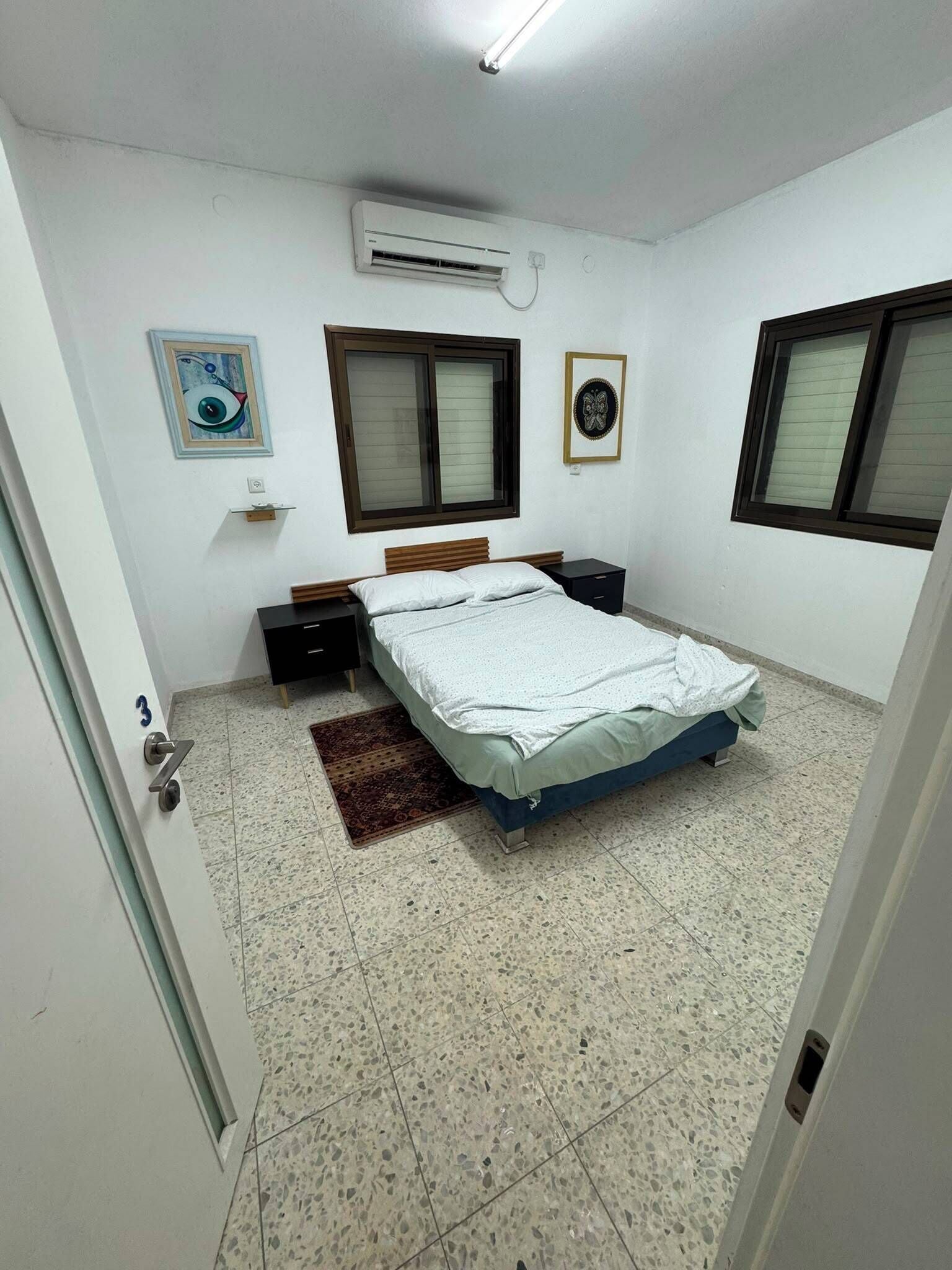 A bedroom with a bed, a nightstand, a wall-mounted air conditioner, a window, and a door.