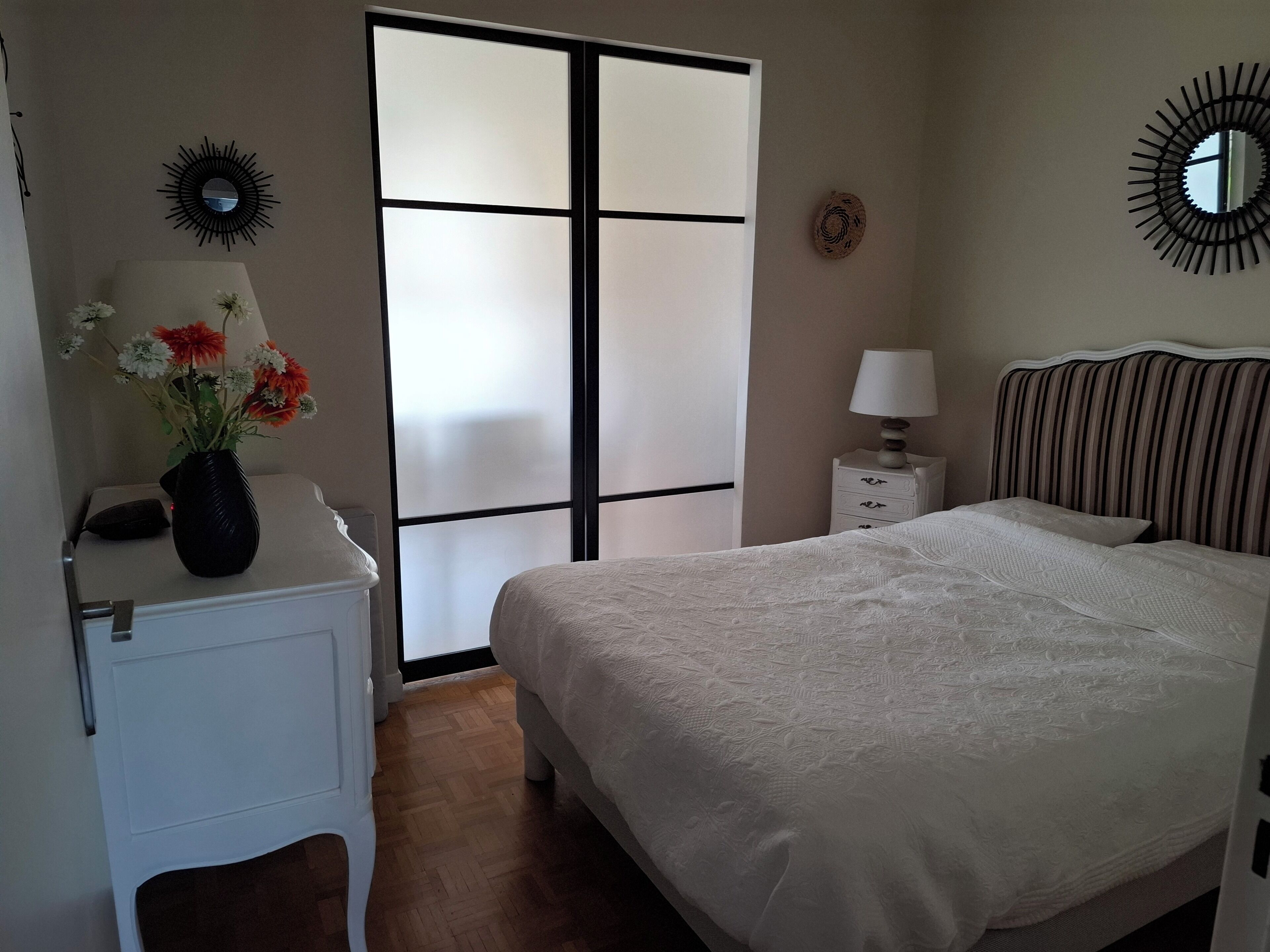 Un dormitorio con una cama, una mesita de noche, una lámpara, un florero con flores, un espejo y una puerta corredera.