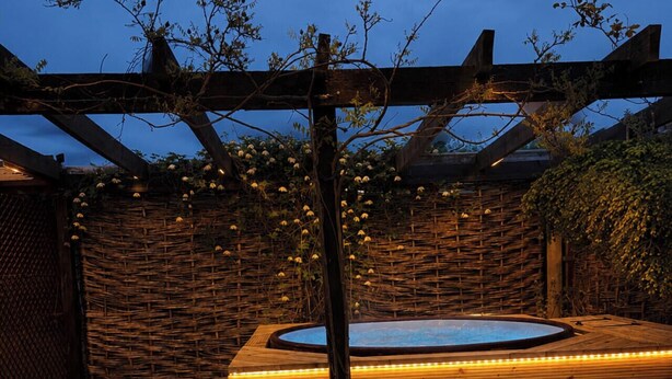Ett trädäck med ett bubbelbad, omgivet av ett trästaket och en pergola med ljusslingor.