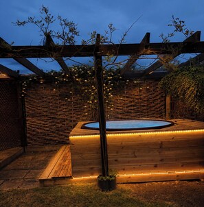 Een houten terras met een jacuzzi, omringd door een houten hek en een pergola met lichtslingers.