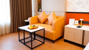 Une chambre d’hôtel moderne avec un canapé, une petite table sur laquelle sont disposés des fruits, une table de chevet avec une lampe et une étagère murale agrémentée d’une plante.