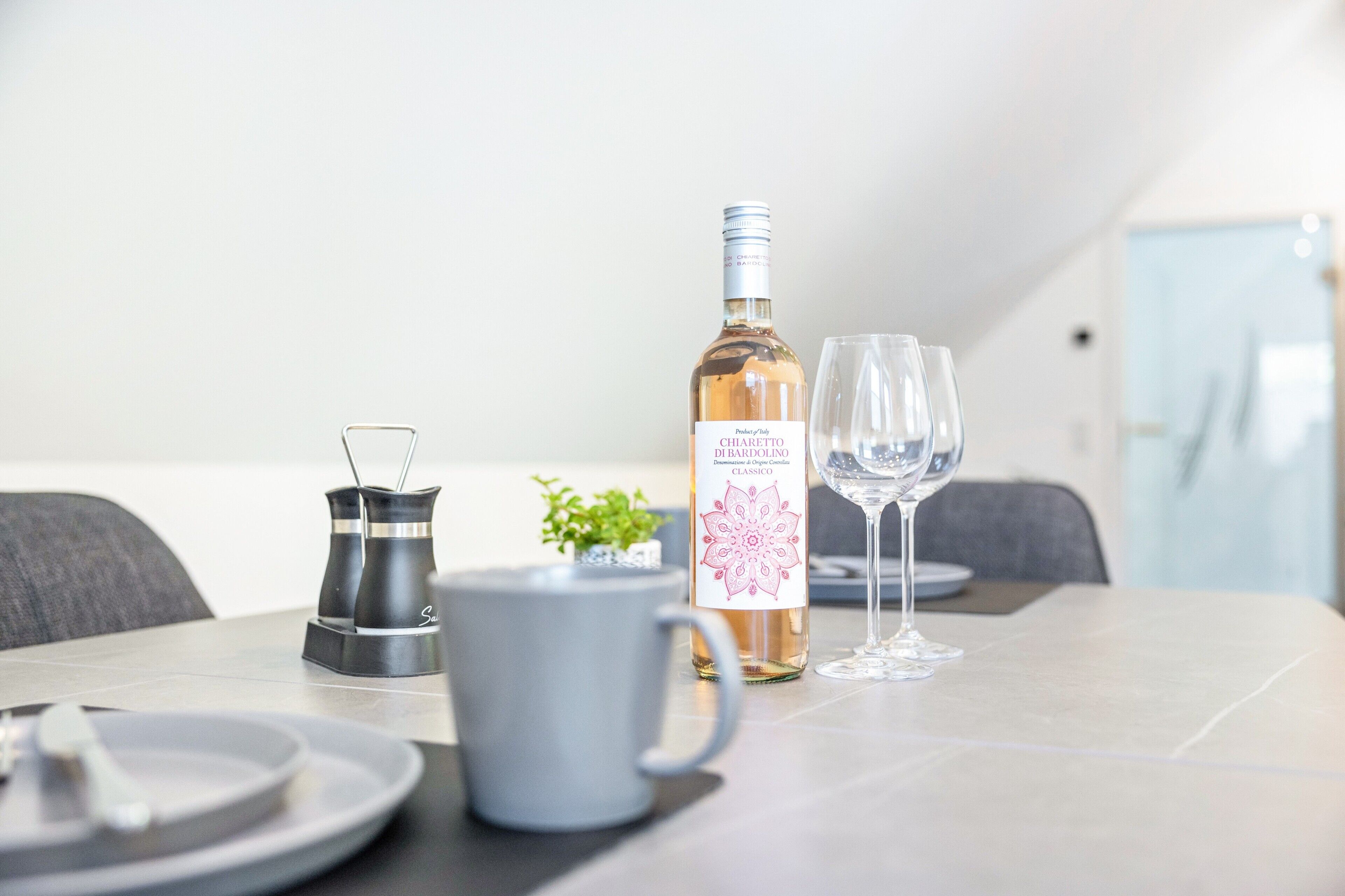 Una mesa de comedor con una botella de vino, dos copas, una cafetera y una planta pequeña.