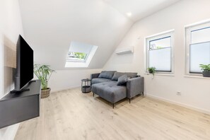Un salon moderne avec un téléviseur à écran plat, un canapé gris, des plantes en pot et une lucarne.
