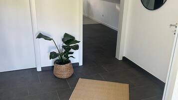Un couloir avec un sol carrelé foncé, un pot de fleurs en fibres tressées, un panneau d’accueil et un miroir rond accroché au mur.