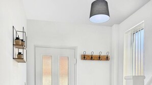 Un couloir intérieur aux murs blancs, avec une porte en verre dépoli, un luminaire suspendu, un porte-manteau et un petit banc en bois.