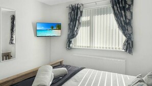 Une chambre à coucher comprenant un lit, un téléviseur, une fenêtre avec des rideaux, un radiateur et un luminaire au plafond.