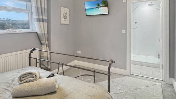 Ein Schlafzimmer mit Bett, Nachttisch, Fernseher, Fenster mit Vorhängen und einer Tür zu einem Duschbereich.