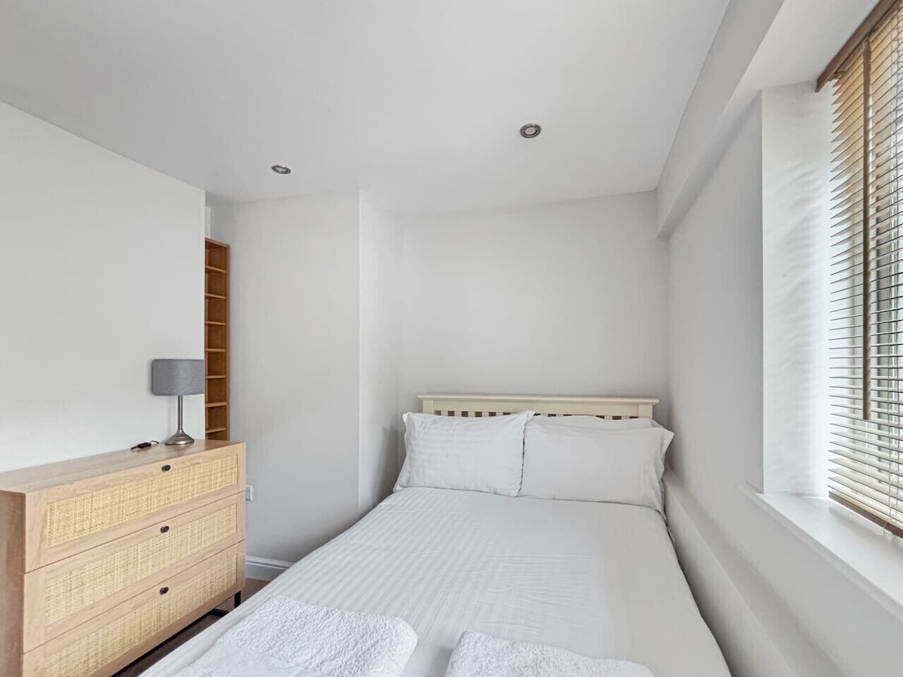Un dormitorio pequeño y ordenado con una cama individual, un aparador de madera, una lámpara y una ventana con persianas.