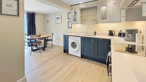 Een kleine keuken met een ingebouwde wasmachine, een eethoek met tafel en stoelen, en een groot raam met gordijnen.