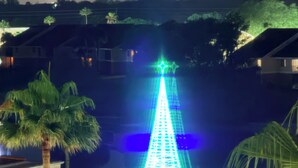 Nachtansicht eines Wasserspiels mit einer sternförmigen Lichtkonstruktion, Palmen und Wohnhäusern im Hintergrund.