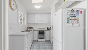 A compact kitchen with white cabinets, a microwave, an oven, and a refrigerator with a calendar. - Bright 3-bed Retreat Near Beaches (Hervey Bay)