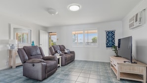 A living room with two reclining sofas, a TV, a small table, and a window with blinds. - Bright 3-bed Retreat Near Beaches (Hervey Bay)