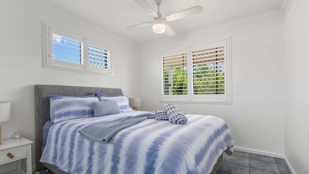 A bedroom with a bed, nightstand, lamp, ceiling fan, and windows with blinds. - Bright 3-bed Retreat Near Beaches (Hervey Bay)