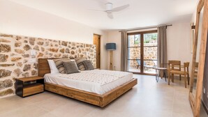 Quarto com parede de pedra, cama de madeira, ventilador de teto e vista através de uma porta de vidro.