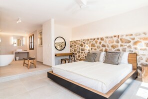A bedroom with a stone wall, a large bed, a wooden nightstand, and a bathroom with a bathtub and vanity. - Luxury Villa Overlooking Sea (PM)