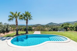 A swimming pool with a curved edge, surrounded by a paved deck, palm trees, and a view of a hilly landscape. - Luxury Villa Overlooking Sea (PM)