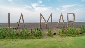 Ein Strand mit Holzbauten, die den Schriftzug 'LAMAR' bilden, und grünen Pflanzen im Vordergrund.