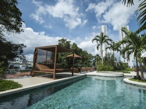 Una piscina con sillas de descanso y una pérgola, rodeada de palmeras y edificios modernos.