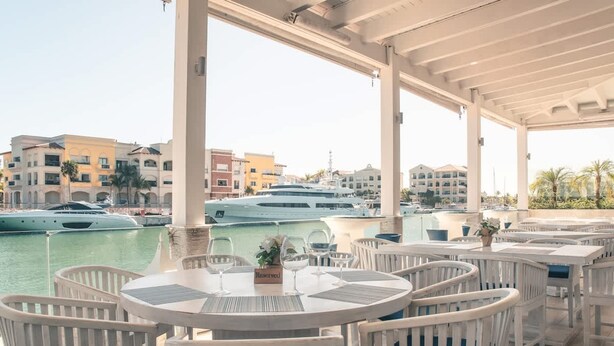 A covered outdoor seating area with white wicker furniture, a view of the marina with yachts, and buildings in the background. - Studio 3 Guests Balcony With Views Fishing Lodge (Punta Cana)