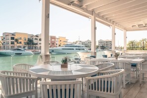 A covered outdoor seating area with white wicker furniture, a view of the marina with yachts, and buildings in the background. - Studio 3 Guests Balcony With Views Fishing Lodge (Punta Cana)