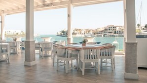 A covered outdoor seating area with white chairs and tables, overlooking a body of water and buildings. - Studio 3 Guests Balcony With Views Fishing Lodge (Punta Cana)