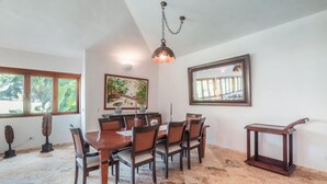A dining area with a wooden table and chairs, a hanging light fixture, a painting, and a window with a view. - 3br Villa Private Pool 6 Guests in Casa de cam (La Romana)