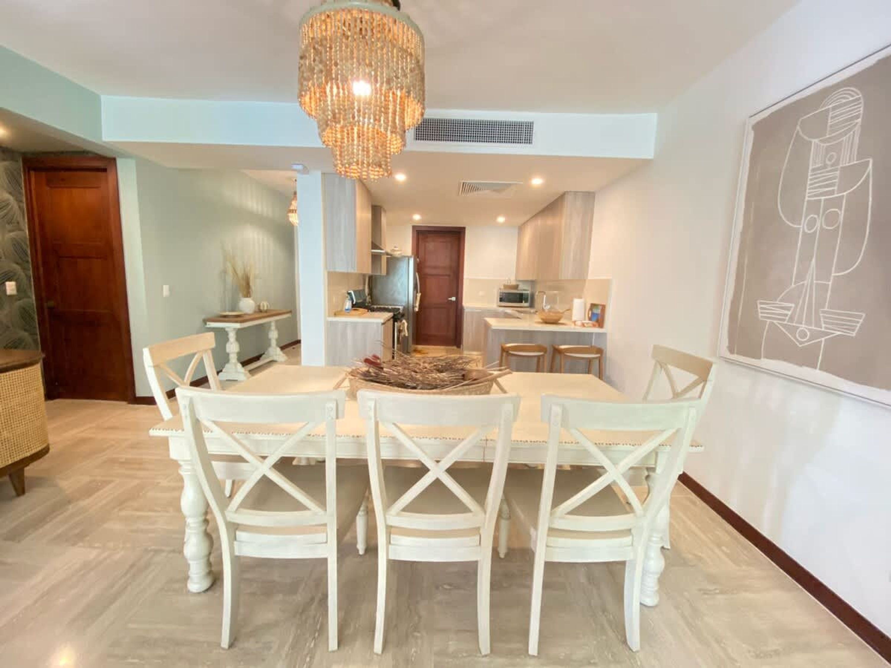 Un comedor con candelabro, sillas blancas, una mesa de madera y una cocina al fondo.