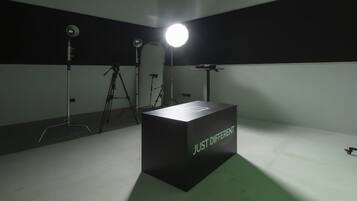 A studio setup with a spotlight, a tripod, and a box labeled 'JUST DIFFERENT' in a room with a plain wall and floor.
