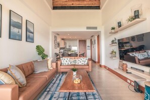 Uma sala de estar moderna com sofá, mesa de centro e televisão. Cozinha visível ao fundo.