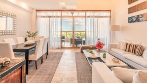 A spacious living room with a dining area, a sofa, a coffee table, and a view through large sliding glass doors to an outdoor patio. - Modern 2br-apartment w Golf View at Casa de Campo (La Romana)